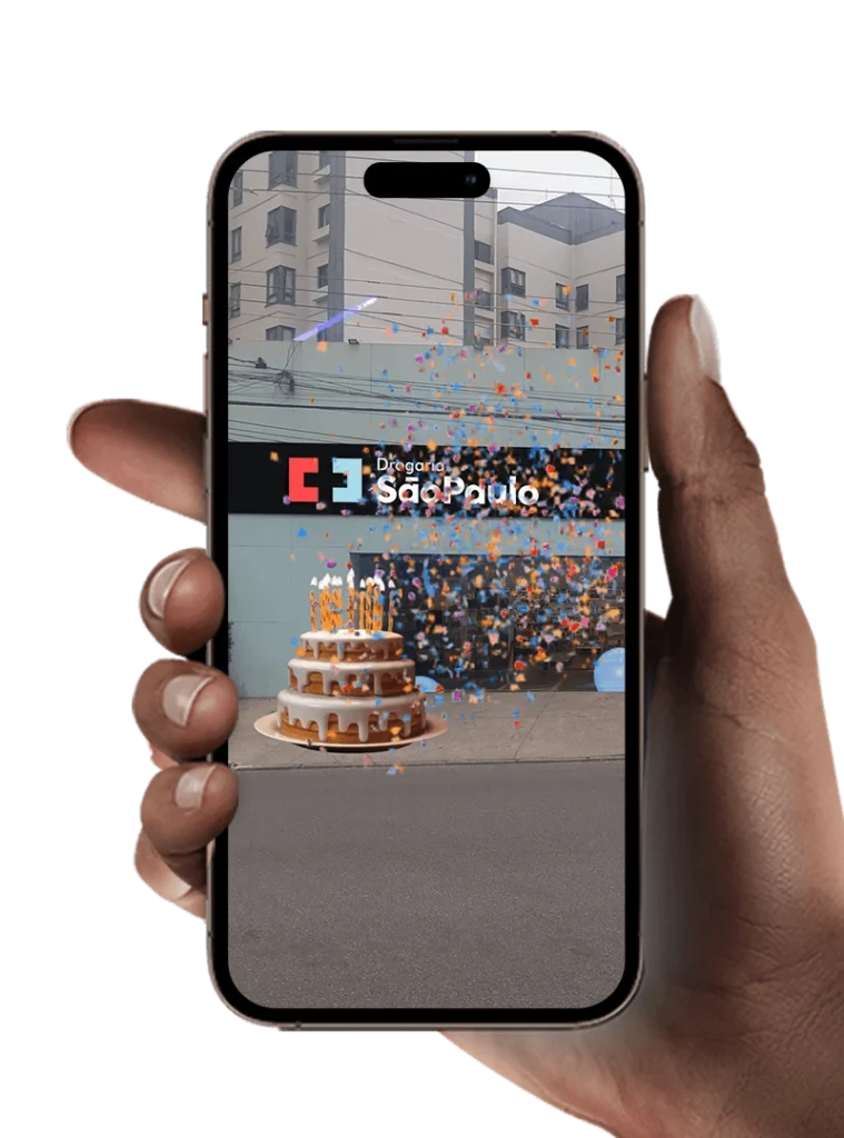 Pessoa segurando celular que mostra um bolo de aniversário em realidade aumentada em frente à fachada da Drogaria São Paulo, com confetes coloridos.