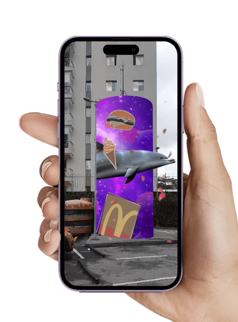 Mão segurando um celular que exibe uma experiência de realidade aumentada com um golfinho nadando em frente a um painel com fundo roxo, hamburguer, sorvete e sacola do McDonald’s flutuando.